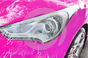 Front headlight of pink car washed in self serve carwash, water spraying leaves white foam on glass