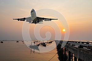Front image commercial passenger aircraft or cargo airplane fly over fishing wood boat floating in the sea at jetty in evening