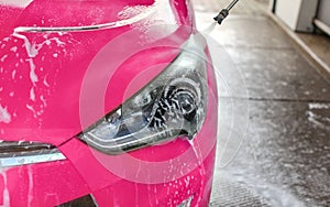 Front light of pink car washed in self serve carwash, jet water spraying on transparent plastics