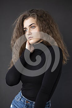 Front photo of a feminity young woman with curly hair, wear black suitcase and jeans, posing near grey wall.