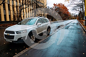 Front side view of white crossover Chevrolet Captiva parked along wet street. Car Chevrolet in autumn landscape