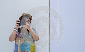 Front view of Asian female tourist photographing with digital camera while leaning against the wall inside of white room