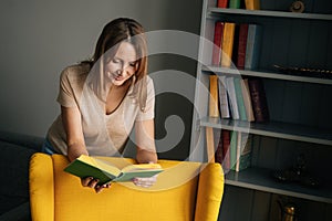 Front view of attractive blonde woman standing near yellow chair reading paper book in dark room with modern interior,