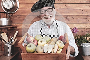 Front view of an attractive and cheerful senior man holding a basket full of apples of various qualities and colors. Rustic
