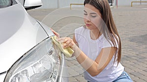 Front view of attractive happy joyful female driver washing her car's headlights with special rag in self-service car