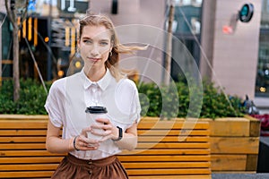 Front view of attractive young woman in casual clothes sitting on bench holding paper cup of takeaway coffee looking at