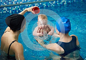 Front view of baby boy during swimming lesson. Back view of mother holding child and woman pouring water on child`s head