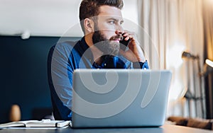 Front view. Bearded businessman is sitting at table in front of computer, talking on cell phone. Entrepreneur works home