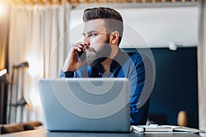 Front view. Bearded businessman is sitting at table in front of computer, talking on cell phone. Entrepreneur works home