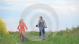 Front view of the beautiful cheerful co uple of lovers actively riding the bicycles along the green blooming field.