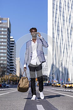 Front view of a black african young man walking on the street wearing elegant jacket and holding a bag while using phone in sunny