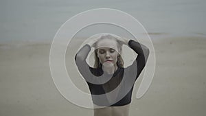 Front view of blonde woman wearing a black elegant swimsuit posing on the beach over sea background - video in slow motion