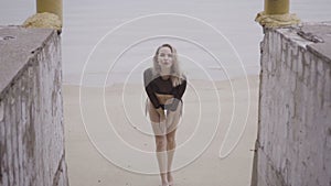 Front view of blonde woman wearing a black elegant swimsuit posing on the beach over sea background - video in slow motion