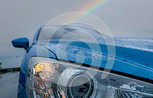 Front view of blue luxury SUV car parked near sea beach on rainy day with rainbow as background. Raindrops on blue texture of car