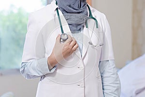 Front view body part of hijab female doctor hand holding stethoscope head inside a hospital room using white lab coat and grey sca