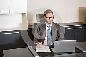 Front view of businessman sitting at table with laptop and holding pen