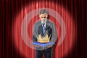 Front view of businessman standing in spotlight against red stage curtain looking down at gold crown on blue pillow