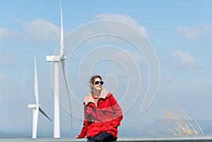 Front view of Caucasian woman with red coat sit on barrier of roadside and use mobile phone on mountain near windmill or wind