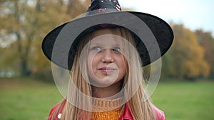 Front view charming cute teen girl in witch hat looking at camera smiling standing outdoors on Halloween. Portrait of