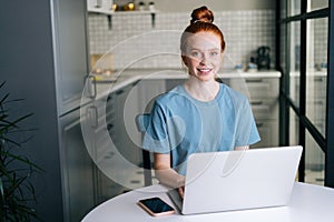 Front view of cheerful redhead young woman working typing on laptop computer sitting at table.