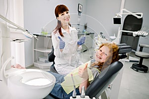 Front view of cheerful smiling female dentist looking at camera with dental tools, and little red haired girl patient