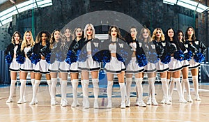 Front view of cheerleaders in uniforms and blue pom-poms standing in v formation in sports hall.