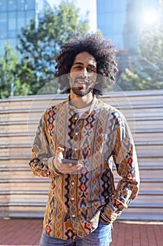 Front view of a cool young smiling afro man using a mobile phone while standing outdoors in a sunny day