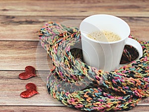 Front view of cup of latte coffee  wrap in colorful scarf and two red glitter hearts on wooden table