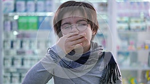 Front view cute boy in eyeglasses sneezing standing in pharmacy indoors. Portrait of Caucasian ill kid waiting for