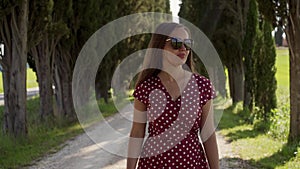 Front view of cute woman with sunglasses in red dress walks among cypress alley