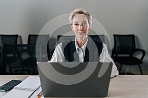Front view of elegance middle-aged businesswoman executive top manager sitting at desk working with documents typing on