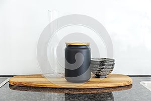 Front view of an elegant wooden tray with a black vase with wooden lid, a glass pitcher and a bowl resting on it