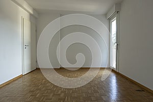 Front view of an empty room with parquet flooring and white walls. On the left is a window letting in a lot of natural light, on