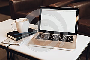 Front view. Empty workplace. On white coffee table is laptop with blank screen, cup of coffee, notebook, pen, newspaper.