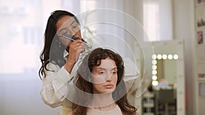 Front view of female hairdresser combing long hair of young woman client with comb after curling with tongs in modern