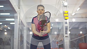 Front view of focused Caucasian sportswoman standing in gym with tennis racket and looking away. Portrait of