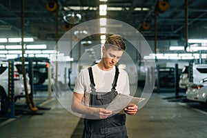 Front view of focused handsome young mechanic male wearing uniform reading clipboard standing in auto repair shop garage