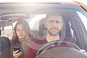 Front view of focused male driver sits at wheel and drives car while his girlfriend sits on back seat, holds smart phone and messa