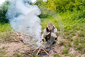 Front view of frozen survivalist male in raincoat putting firewood on campfire to making fire on bank of river in photo