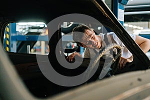 Front view of frustrated young service man in uniform inspecting interior of old car in auto repair shop garage with