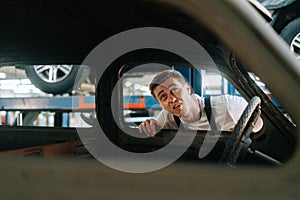 Front view of frustrated young service man in uniform inspecting interior of old car in auto repair shop garage with