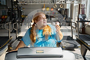 Front view on funny obese woman eating cake while training on treadmill at gym
