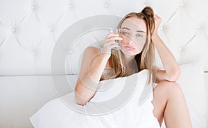 Front view of a girl with tousled hair hold glass water, suffering head ache sitting on bed in the living room at home.Woman hango