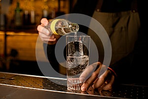front view of glass with cold beverage and ice cubes into which bartender accurate pours drink from bottle