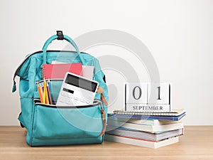 Front view of green backpack with school supplies, stack of books and wooden calendar September 01 on wooden table and white