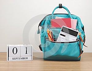 Front view of green backpack with school supplies and wooden calendar September 01 on wooden table and white background with