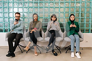 Front view of group young diverse ethnicity candidates using mobile phones while waiting job interview in modern office