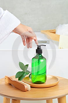 Front view of a hand above an unlabeled shampoo bottle with spray head displayed on a wooden plate, next to a green leaf branch