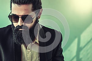 Front view of handsome bearded man with a man bun hairstyle posing in studio wearing a suit and sunglasses on green background,