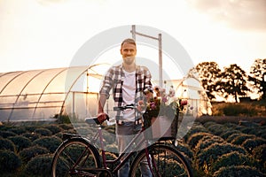 Front view. Handsome man in casual clothes is with bicycle on the agricultural field near greenhouse